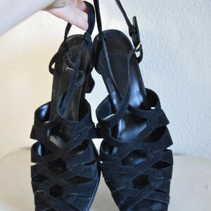 May include: A pair of black suede sandals with multiple straps and a slingback design. The sandals have a small heel and a buckle closure. The shoes are held up by a hand.