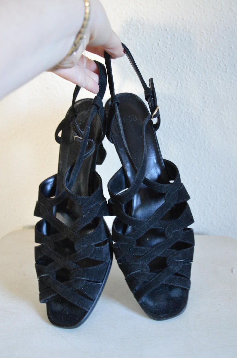 May include: A pair of black suede sandals with multiple straps and a slingback design. The sandals have a small heel and a buckle closure. The shoes are held up by a hand.