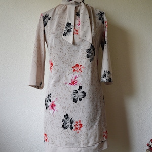 Può includere: Un vestito beige con stampa floreale nera e rossa e un fiocco bianco al collo.