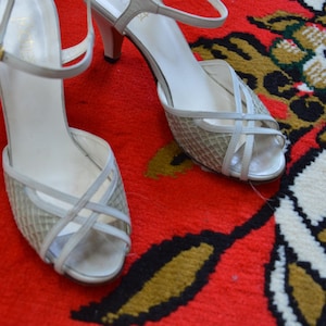 Puede incluir: Un par de sandalias de tac&oacute;n color blanco roto con un dise&ntilde;o de tiras cruzadas sobre la puntera abierta y una tira fina en el tobillo con una hebilla dorada. La parte delantera del zapato est&aacute; hecha de un material de malla tejida texturizada.