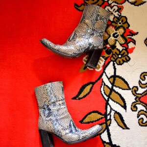 May include: Two ankle boots with a snakeskin print in grey, brown, and blue tones. The boots feature a pointed toe and a block heel. The boots are placed on a red and cream patterned surface.