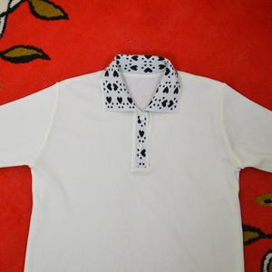 Pode incluir: Camisa polo creme com gola e carcela com botões com padrão de corações em preto e branco. A camisa tem mangas curtas e um design clássico de polo.