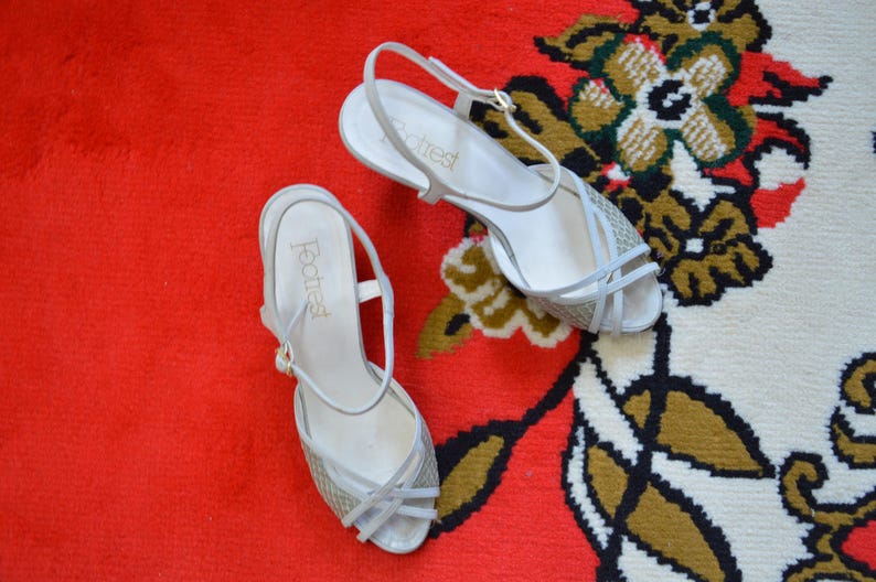 Puede incluir: Un par de sandalias blancas con tira trasera y cierre de hebilla. La parte delantera de las sandalias presenta un patr&oacute;n texturizado tipo tejido y tiras finas. El nombre de la marca Footrest est&aacute; impreso en letras doradas en la plantilla blanca.