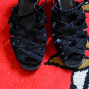 May include: A pair of black suede sandals with multiple crisscrossing straps. The open-toe shoes have a small heel and ankle straps. The sandals are displayed on a red and patterned surface.