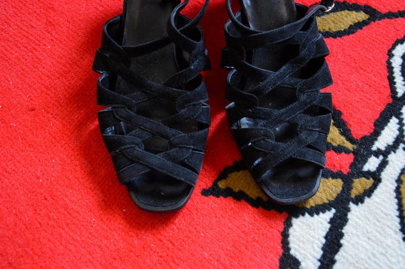May include: A pair of black suede sandals with multiple crisscrossing straps. The open-toe shoes have a small heel and ankle straps. The sandals are displayed on a red and patterned surface.