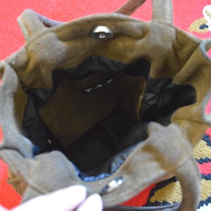 May include: A brown suede tote bag with a structured design. The bag has a top handle and a magnetic snap closure. The interior is black, and a small pocket is visible. The bag is sitting on a red and patterned surface.