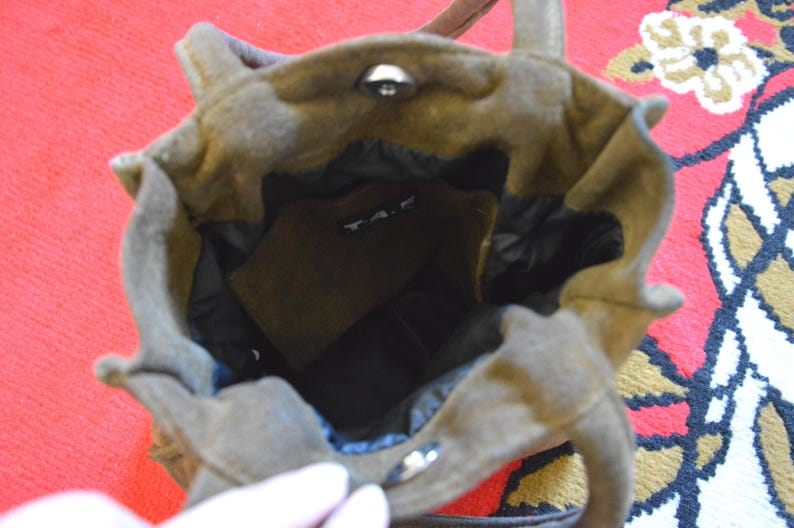 May include: A brown suede tote bag with a structured design. The bag has a top handle and a magnetic snap closure. The interior is black, and a small pocket is visible. The bag is sitting on a red and patterned surface.