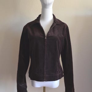 May include: A dark brown, zip-up jacket made of a soft, velvety material. The jacket has a collar and long sleeves. It is displayed on a mannequin against a neutral background. The jacket is a medium size.