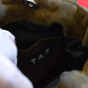 May include: Close-up of a brown suede bag with a black interior. The bag has a magnetic closure and a label that reads "T.A.F." in white letters. The bag is open, revealing the inside.