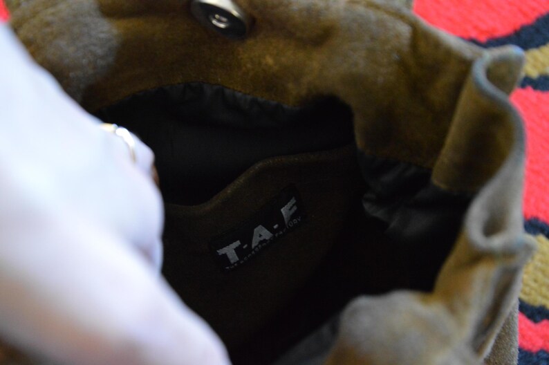 May include: Close-up of a brown suede bag with a black interior. The bag has a magnetic closure and a label that reads "T.A.F." in white letters. The bag is open, revealing the inside.