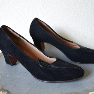 May include: A pair of black suede heeled shoes with a pointed toe and a small black leather leaf detail on the vamp.