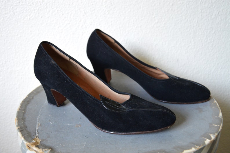 May include: A pair of black suede heeled shoes with a pointed toe and a small black leather leaf detail on the vamp.