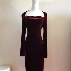 May include: A burgundy velvet dress with long sleeves and a square neckline. The dress is displayed on a mannequin and has a fitted silhouette. The fabric appears soft and drapes smoothly. The dress is knee-length.
