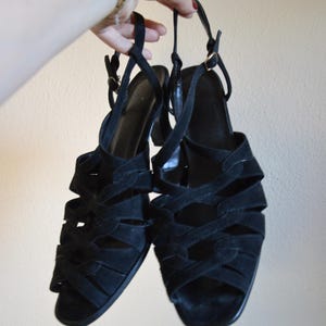 May include: A pair of black suede sandals with multiple straps and a small heel. The sandals have a strappy design and are held up by a person's hand. The sandals have a buckle closure and are a classic style.