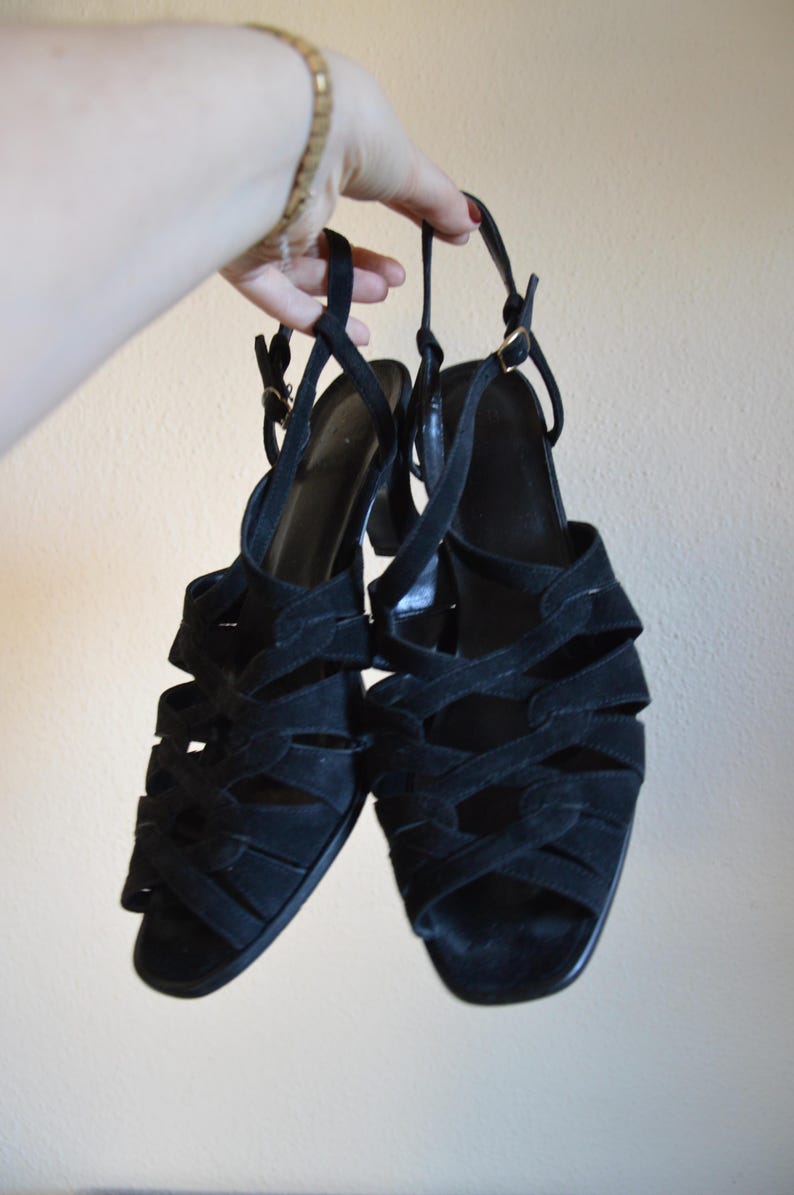 May include: A pair of black suede sandals with multiple straps and a small heel. The sandals have a strappy design and are held up by a person's hand. The sandals have a buckle closure and are a classic style.