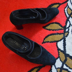 May include: A pair of black suede shoes with a low heel and a strap across the top. The shoes are on a red and white patterned surface. The shoes are labeled "Footrest".