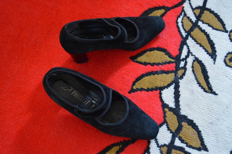 May include: A pair of black suede shoes with a low heel and a strap across the top. The shoes are on a red and white patterned surface. The shoes are labeled "Footrest".