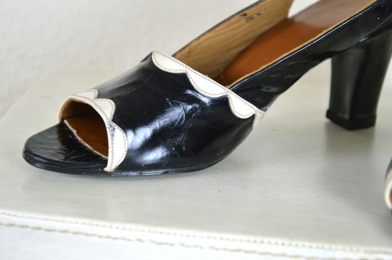 May include: Black leather heeled shoes with a white scalloped trim around the opening. The shoes have a closed toe and a peep toe design.