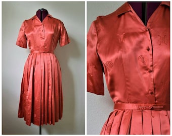 May include: A coral red satin dress with a button-up front and a pleated skirt. The dress has a short sleeve and a collar. The fabric has a subtle embroidered pattern.