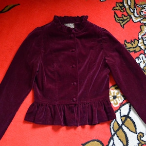 May include: A deep burgundy velvet jacket featuring a high ruffled collar, long sleeves, and a peplum hem. The front closure consists of a vertical row of matching fabric-covered buttons. A small rectangular label is visible at the inner neckline.