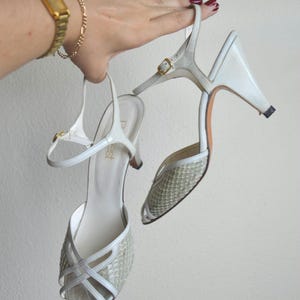 Puede incluir: Un par de zapatos de tac&oacute;n blancos con tira trasera, que presentan una puntera texturizada con patr&oacute;n de piel de serpiente y tiras cruzadas. Los zapatos tienen una hebilla dorada en la tira del tobillo y un tac&oacute;n medio. La plantilla muestra la marca 'Dest'.