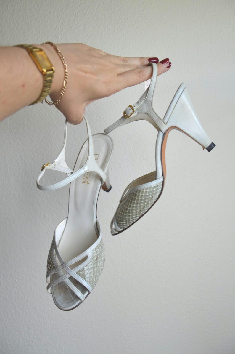 Puede incluir: Un par de zapatos de tac&oacute;n blancos con tira trasera, que presentan una puntera texturizada con patr&oacute;n de piel de serpiente y tiras cruzadas. Los zapatos tienen una hebilla dorada en la tira del tobillo y un tac&oacute;n medio. La plantilla muestra la marca 'Dest'.