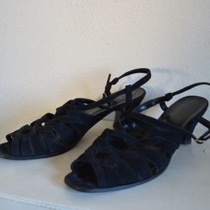 May include: A pair of black suede sandals with open toes and a strappy design. The sandals have a small heel and adjustable ankle straps. The shoes are displayed on a white surface.