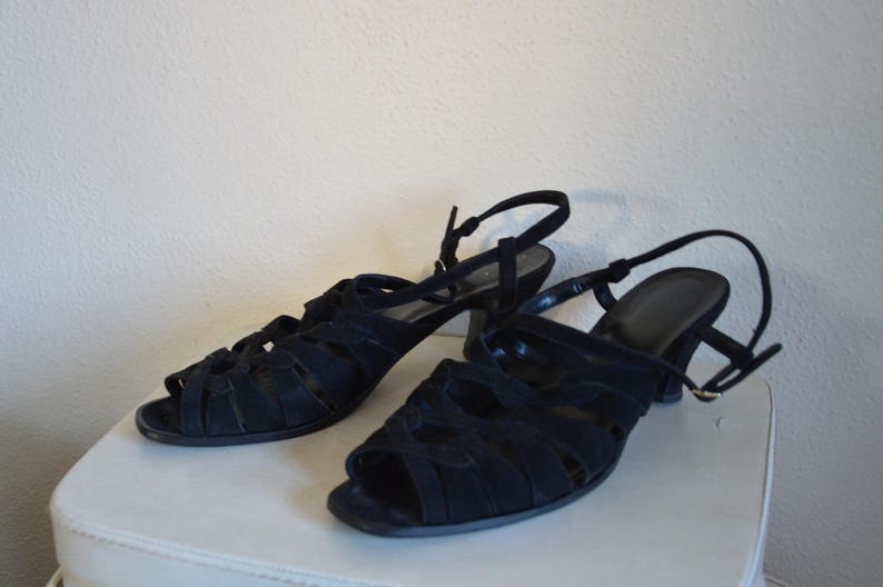 May include: A pair of black suede sandals with open toes and a strappy design. The sandals have a small heel and adjustable ankle straps. The shoes are displayed on a white surface.