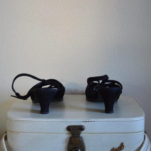 May include: A pair of black, open-toe sandals with small heels. The shoes have a strap that wraps around the ankle. They are placed on top of a white suitcase with a metal clasp.