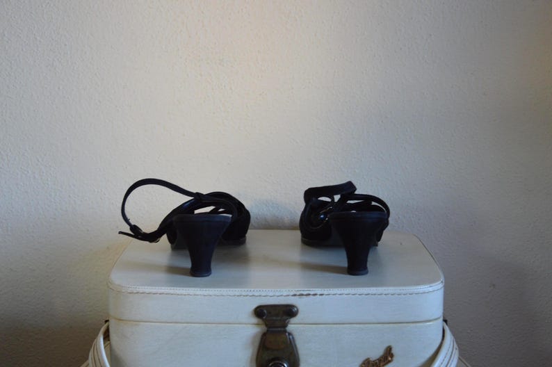 May include: A pair of black, open-toe sandals with small heels. The shoes have a strap that wraps around the ankle. They are placed on top of a white suitcase with a metal clasp.