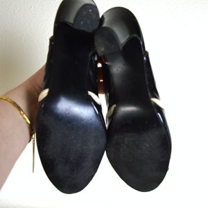 May include: A pair of black high-heeled shoes with white accents. The shoes are made of leather and have a pointed toe.