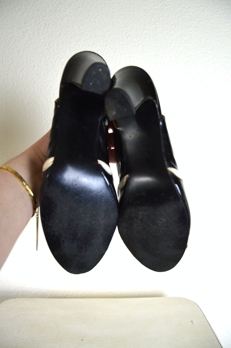 May include: A pair of black high-heeled shoes with white accents. The shoes are made of leather and have a pointed toe.