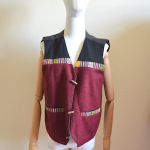 Peut inclure: Un gilet bordeaux avec un col noir et une garniture tissée colorée. Le gilet a deux fermetures à bascule en bois et deux poches avec une garniture assortie. Le gilet est présenté sur un mannequin.