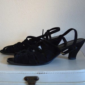 May include: A pair of black suede sandals with open toes and a strappy design. The sandals have a small heel and adjustable ankle straps. The shoes are displayed on a white surface.