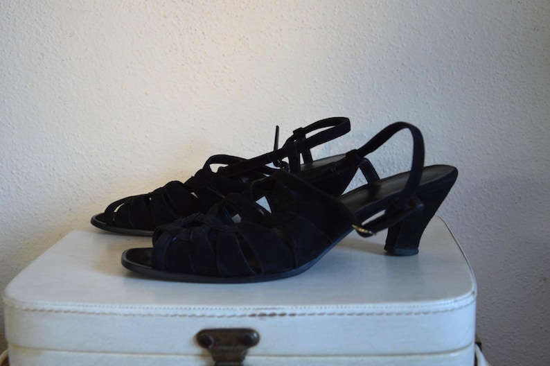 May include: A pair of black suede sandals with open toes and a strappy design. The sandals have a small heel and adjustable ankle straps. The shoes are displayed on a white surface.