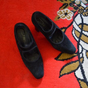 May include: A pair of black suede Mary Jane style shoes with a low heel. The shoes have a strap across the top of the foot and are set against a red background.
