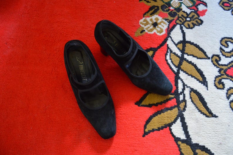 May include: A pair of black suede Mary Jane style shoes with a low heel. The shoes have a strap across the top of the foot and are set against a red background.