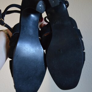 May include: A pair of black suede sandals with a strappy design. The sandals have a small heel and a buckle closure. The soles are black and show some wear. The sandals are held up against a neutral background.