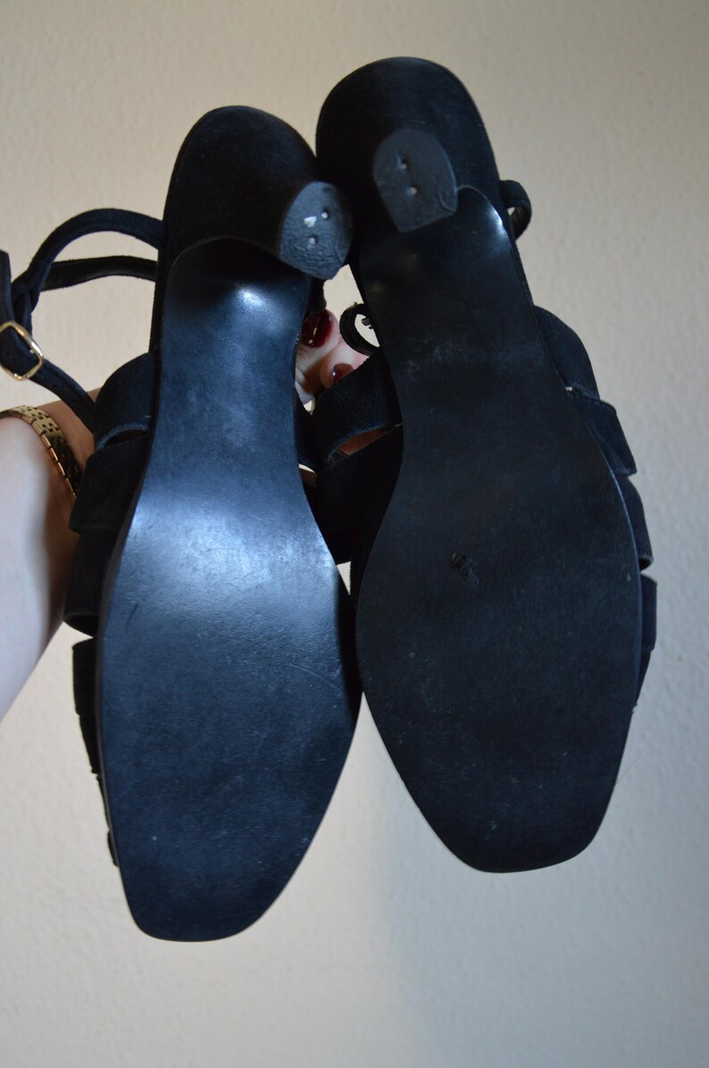 May include: A pair of black suede sandals with a strappy design. The sandals have a small heel and a buckle closure. The soles are black and show some wear. The sandals are held up against a neutral background.