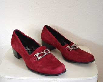 Könnte beinhalten: Ein Paar bordeauxfarbene Loafer mit Absatz aus Wildleder. Jeder Schuh hat eine silberfarbene Metallschnalle und einen schwarzen Absatz. Die Schuhe werden auf einer weißen Oberfläche vor einem weißen Hintergrund präsentiert.