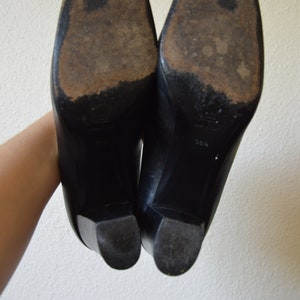 Puede incluir: Un par de zapatos de tac&oacute;n de cuero negro con punta puntiaguda. Los zapatos tienen suela de goma y se muestran desde la parte inferior.