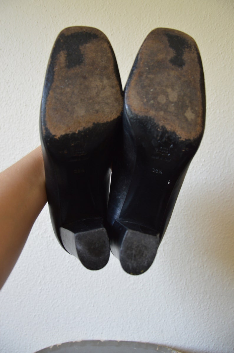Puede incluir: Un par de zapatos de tac&oacute;n de cuero negro con punta puntiaguda. Los zapatos tienen suela de goma y se muestran desde la parte inferior.