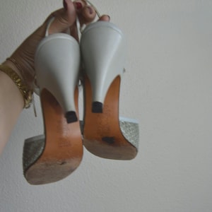 Puede incluir: Un par de zapatos de tac&oacute;n blancos con tira trasera y un patr&oacute;n texturizado en la parte delantera. Los zapatos cuentan con un tac&oacute;n blanco liso, una suela de color tostado y una peque&ntilde;a tapa negra en el tac&oacute;n. Incluyen una tira fina.