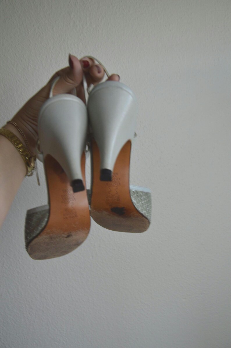 Puede incluir: Un par de zapatos de tac&oacute;n blancos con tira trasera y un patr&oacute;n texturizado en la parte delantera. Los zapatos cuentan con un tac&oacute;n blanco liso, una suela de color tostado y una peque&ntilde;a tapa negra en el tac&oacute;n. Incluyen una tira fina.