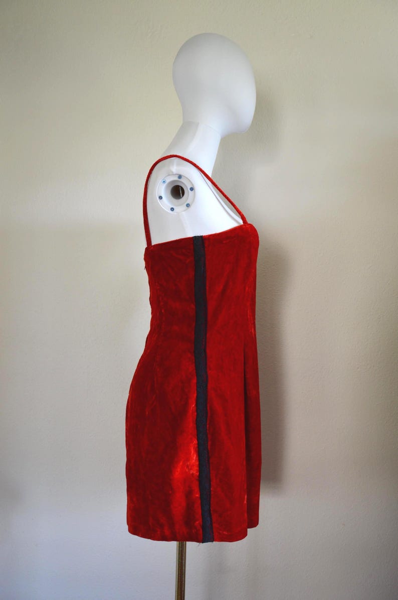 May include: A red velvet mini-dress with thin straps and a dark blue vertical stripe. The dress is displayed on a mannequin. The dress is a classic, simple design.