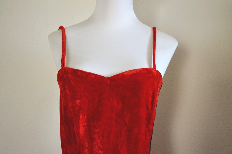 May include: A red velvet dress with thin spaghetti straps. The dress has a sweetheart neckline and is displayed on a white mannequin. The fabric appears soft and luxurious, with a slight sheen.