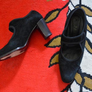 May include: A pair of black suede heeled shoes with a Mary Jane strap design. The shoes have a rounded toe and a block heel. The shoes are displayed on a red rug with a floral pattern.