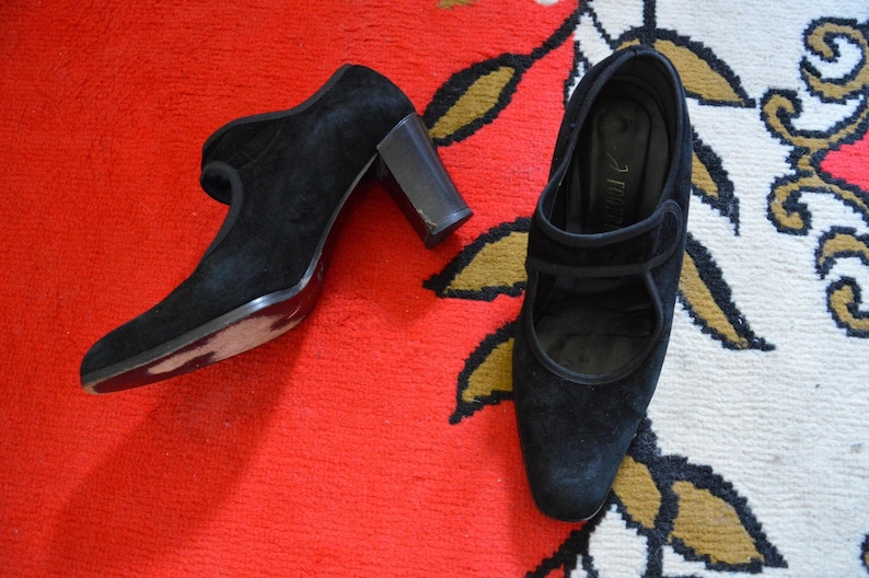 May include: A pair of black suede heeled shoes with a Mary Jane strap design. The shoes have a rounded toe and a block heel. The shoes are displayed on a red rug with a floral pattern.