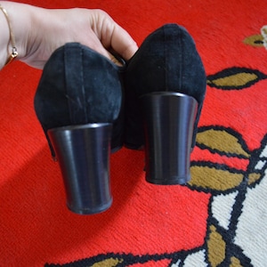 May include: Pair of black suede heels with dark brown block heels. The shoes are held up against a red and patterned background. The heels appear to be approximately 3 inches tall.