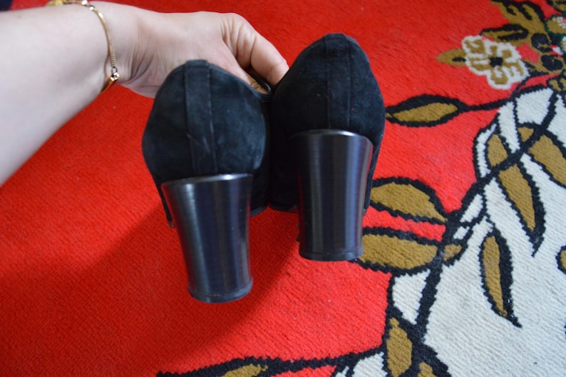 May include: Pair of black suede heels with dark brown block heels. The shoes are held up against a red and patterned background. The heels appear to be approximately 3 inches tall.